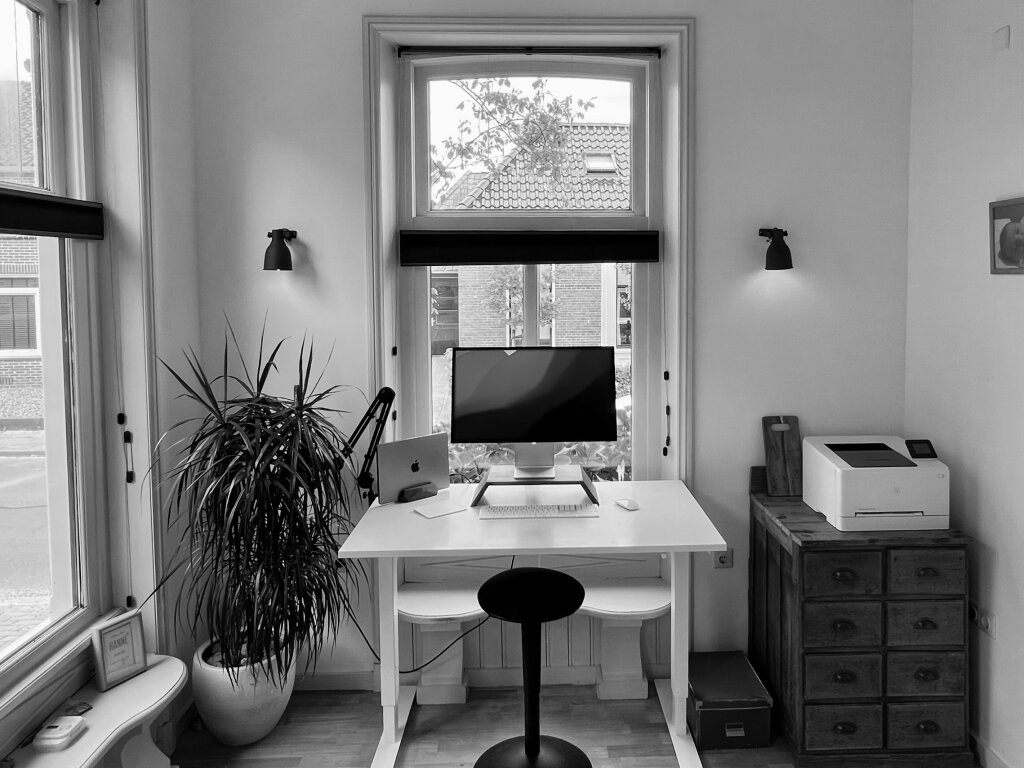 Mijn kantoor, met plant, bureau, scherm, Macbook, printer en een houten vloer en uitzicht naar buiten.